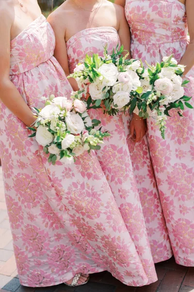 Bridesmaid dresses in pink floral strapless styles, holding white rose and blush ranunculus bouquets with eucalyptus on a brick walkway