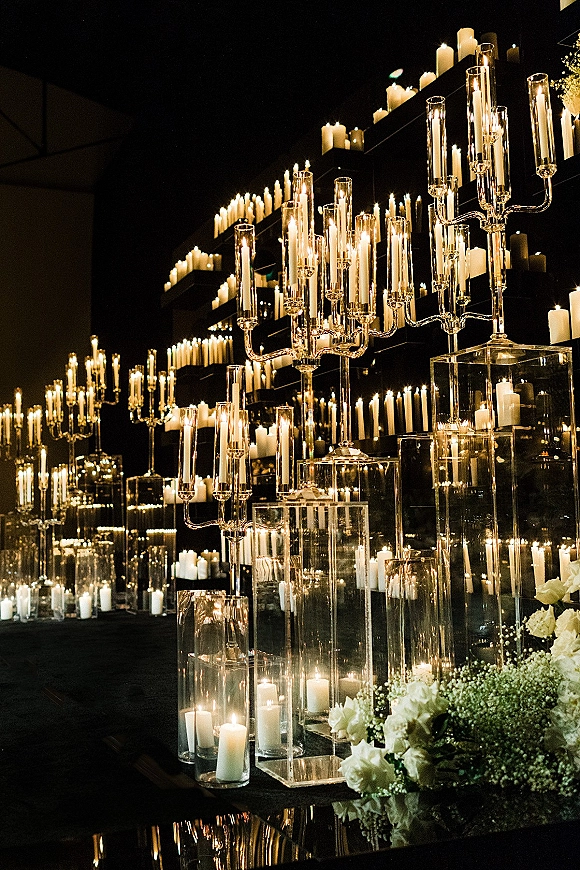 Wedding candle decor with pillar and taper candles in glass candelabras on acrylic pedestals, with white flowers against a dark venue backdrop