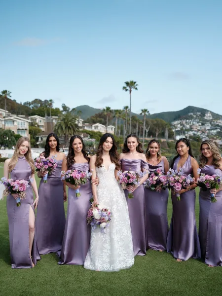 Bridesmaid group portrait with bride and bridesmaids holding bouquets in satin purple dresses on a lawn with palm trees and hills