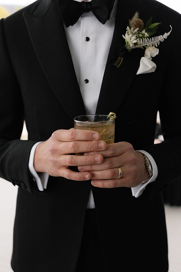 Groom portrait in black tie groom tux, holding a cocktail with lemon twist, showing boutonniere, pocket square, and wedding band on an outdoor walkway