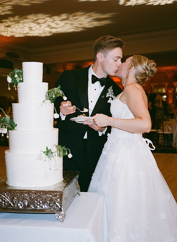 Cake cutting moment as bride and groom kiss beside a tiered wedding cake with greenery on a silver stand in a warmly lit ballroom reception