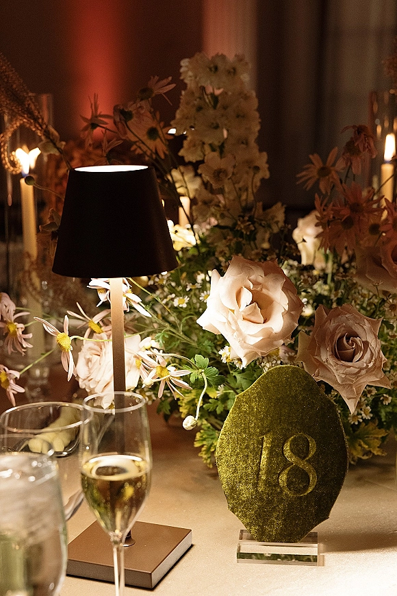 Reception tablescape with wedding table centerpiece of blush roses and white daisies, taper candles, and a table lamp in dim lighting