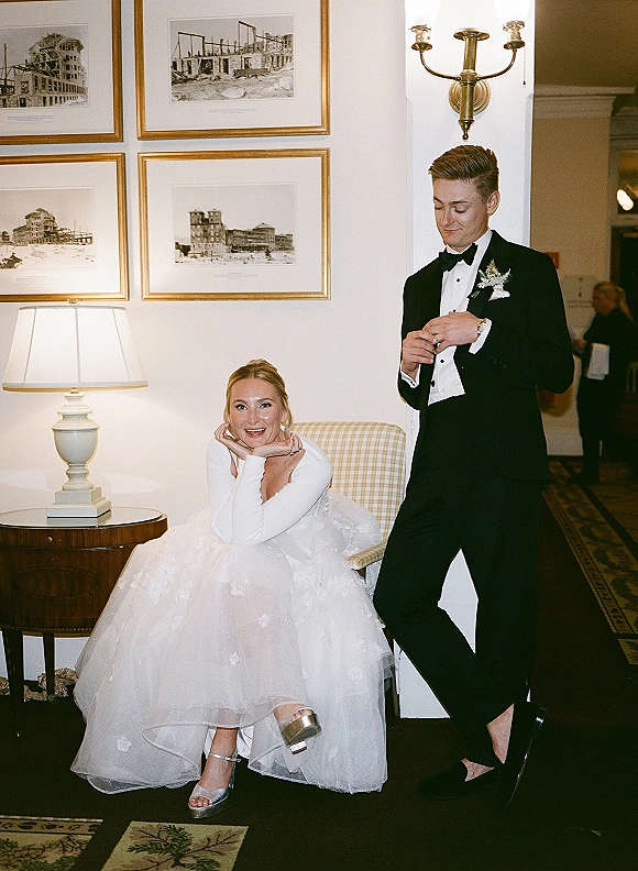 Couple portrait in an indoor wedding portrait setting, bride in long sleeve gown seated as groom in tux stands in a framed hallway