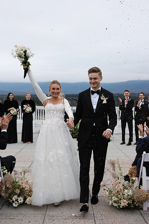 Wedding recessional as bride and groom walk down aisle, bride lifting bouquet overhead while guests clap on a mountain terrace backdrop