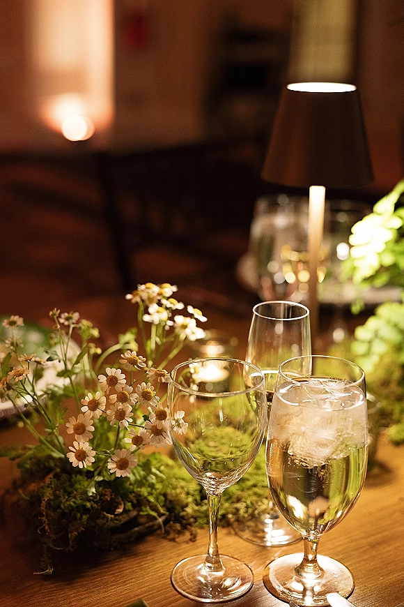 Reception tablescape with a moss table runner, daisies, votive candles, and glassware on a wood table in warm, dim indoor light