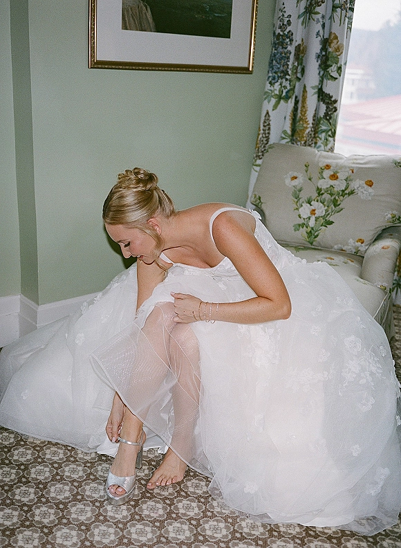 Bride getting ready as she adjusts an ankle strap heel, showing sheer hosiery and tulle dress with floral appliqués by window light