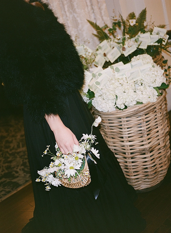 Bridal bouquet of daisies and white hydrangeas in a wicker basket with ribbon, held beside a black dress near a stone wall indoors