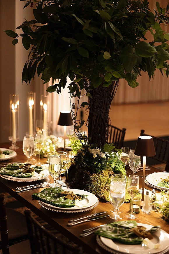 Reception tablescape with green wedding tablescape greenery, tall branch centerpieces, moss runner, taper candles and lamps on a wooden table indoors