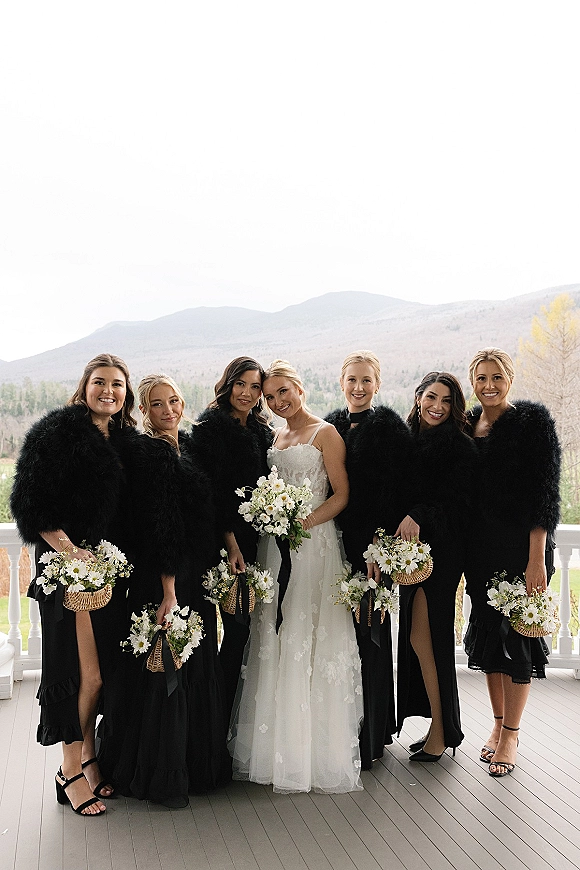 Bridesmaid group photo with bride with bridesmaids in black dresses holding white bouquets and wicker baskets on a porch with mountain views