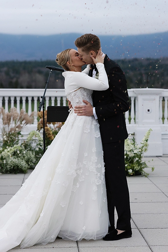 Wedding kiss as confetti falls around bride in lace long-sleeve topper and groom in tux on an outdoor terrace with mountain view