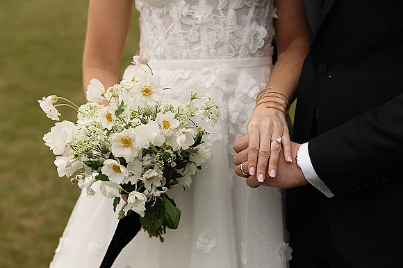 Wedding couple portrait with bride and groom holding hands, engagement ring visible beside a white wildflower bouquet on a green lawn