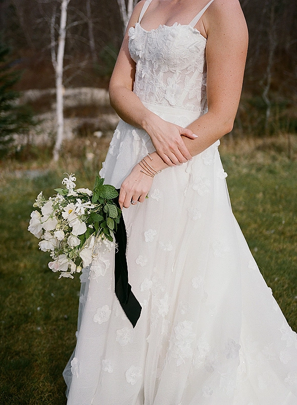 Bridal portrait of a bride in a lace wedding dress holding a white bouquet with greenery and black ribbon on a grassy lawn with trees