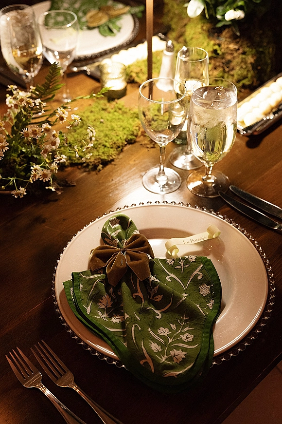 Reception tablescape with wedding place setting, green patterned napkin tied in a velvet bow beside beaded charger, candles and moss runner on wood table