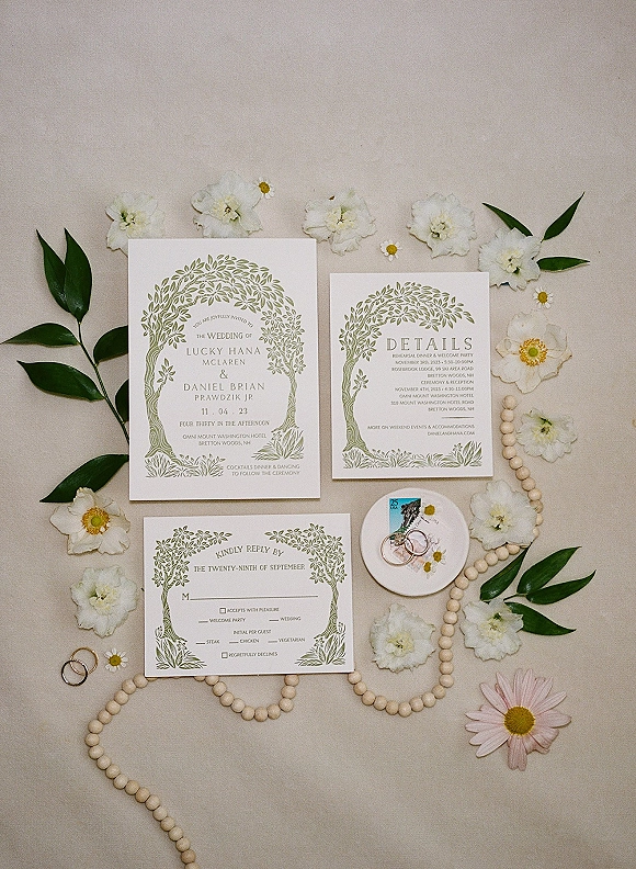 Wedding invitation suite with green botanical wedding invitation cards, rings in a dish, postage stamp, and beaded garland on linen backdrop