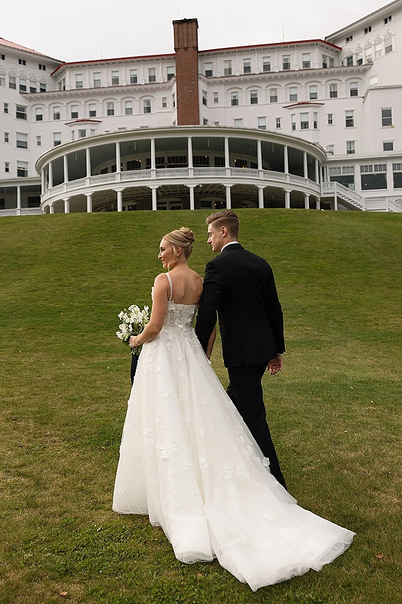 Couple portrait of bride and groom walking hand in hand, bride looking back holding bouquet, with grand hotel veranda behind