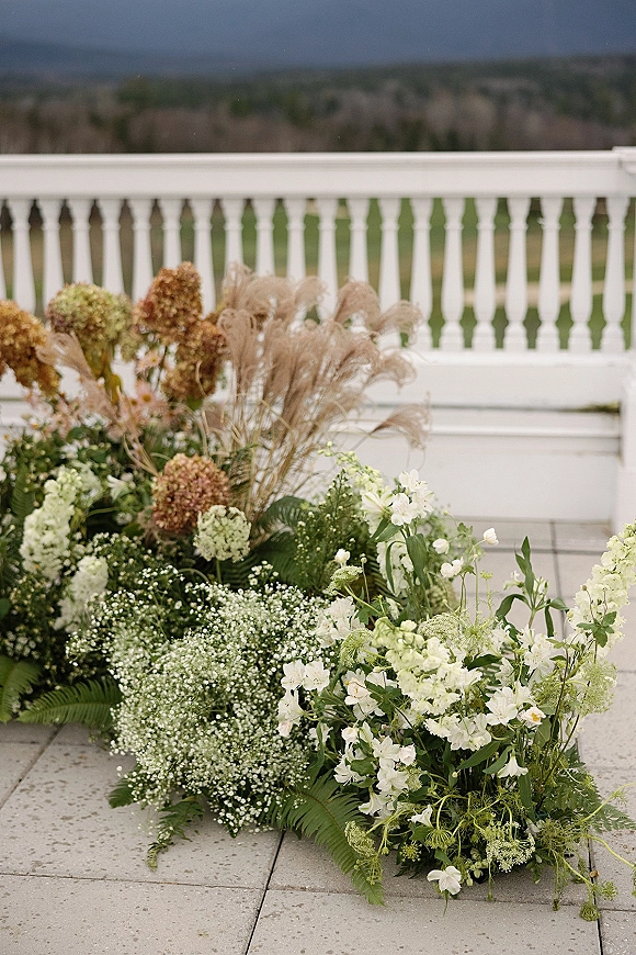 Wedding floral arrangement of white hydrangeas and baby’s breath with greenery and pampas grass by a terrace railing with distant hills