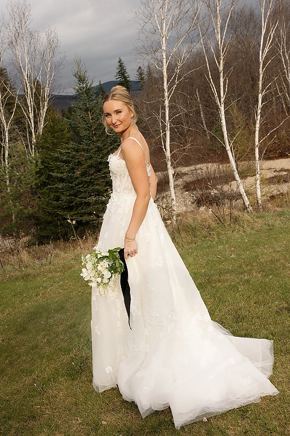 Bridal portrait of a bride holding bouquet of white flowers and greenery, showing a lace dress with train in a mountain meadow under clouds