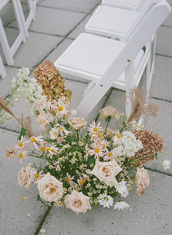 Aisle florals with wedding aisle flowers in blush roses, daisies, and hydrangea arranged beside white folding chairs on stone pavers