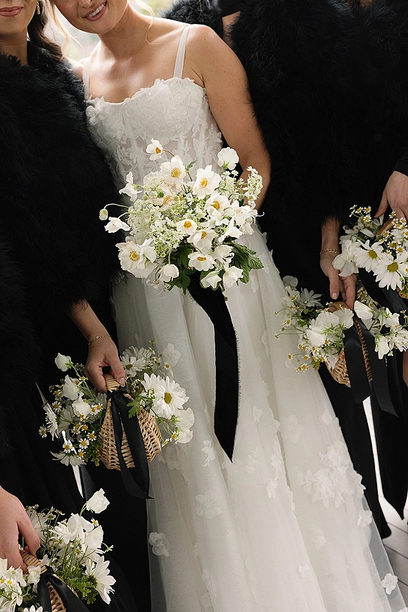 Bride with bridesmaids posing outdoors in greenery, holding daisy wedding bouquet and wicker baskets with black faux fur wraps