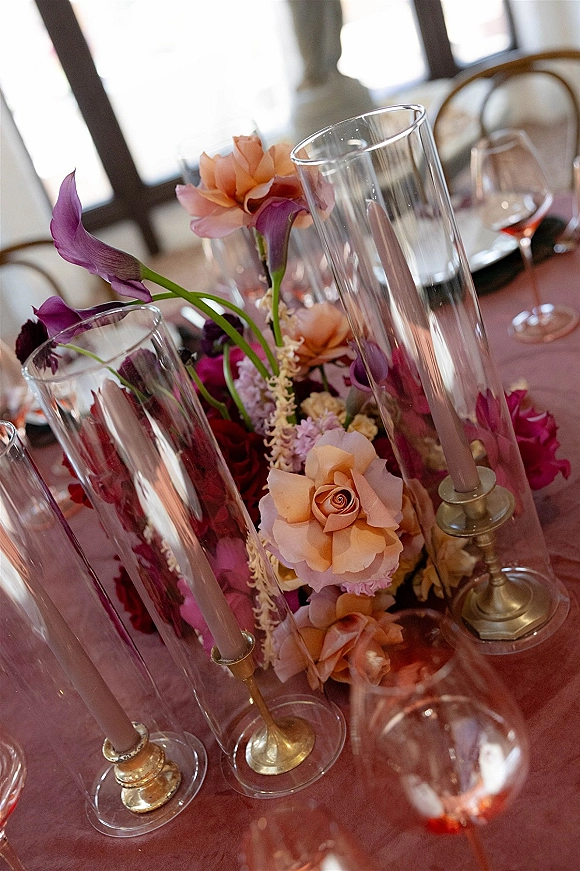 Wedding centerpiece with floral centerpiece candles, roses and calla lilies among glass cylinders, brass candlesticks, and window-lit reception tables