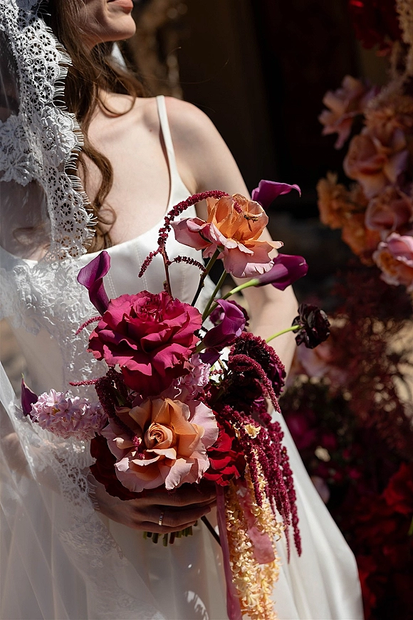 Bridal bouquet with a burgundy wedding bouquet of roses, calla lilies, and hanging amaranthus, held against a dark floral backdrop