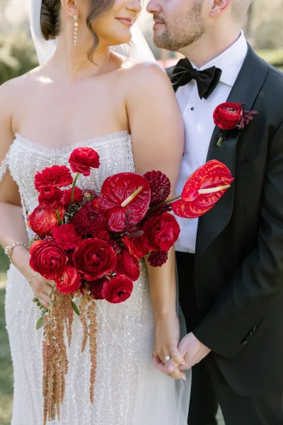Couple portrait of bride and groom close up holding hands, her red bouquet with anthurium, in natural light with blurred greenery behind