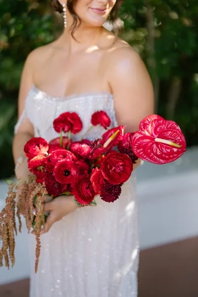 Bridal bouquet of red anthurium and ranunculus held against a strapless lace wedding dress, with pearl drop earrings in a sunlit garden