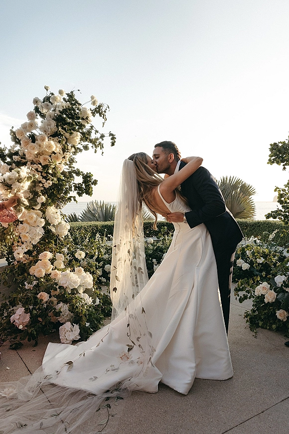 Wedding kiss portrait of bride and groom kiss in a dip beneath a rose and greenery floral arch, veil flowing with ocean view behind