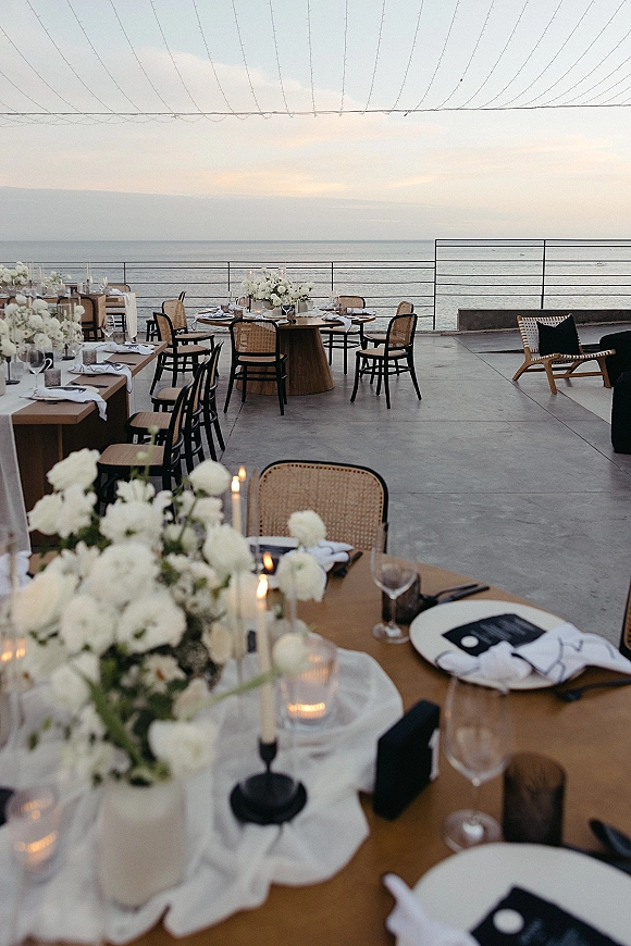 Reception tablescape at an oceanfront wedding reception with white florals, taper and votive candles, cane chairs, and string lights at sunset