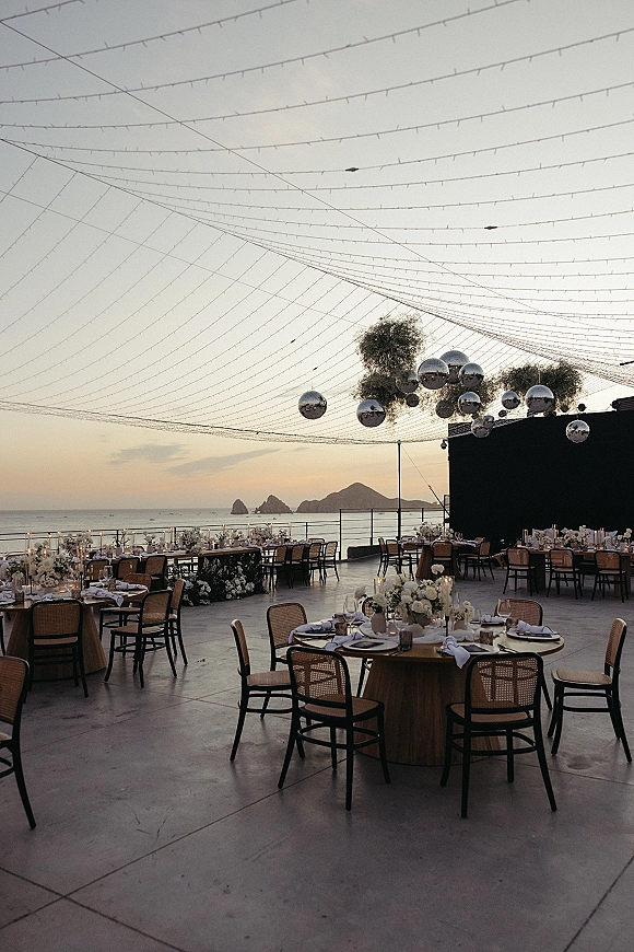 Reception tablescape with round tables, candles and white floral centerpieces under string lights and disco balls on an oceanfront terrace at sunset