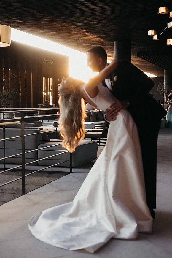 Wedding kiss portrait of groom dipping bride in a long-train dress, backlit at sunset in a modern lobby with pendant lights