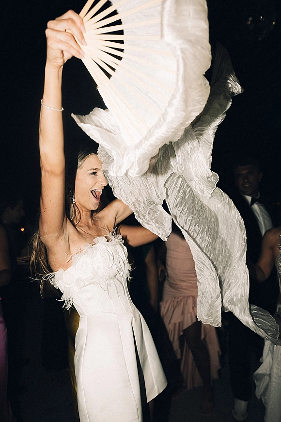 Wedding dance moment as the bride dances reception holding a paper hand fan in a strapless feather-trim mini dress on a dark dance floor