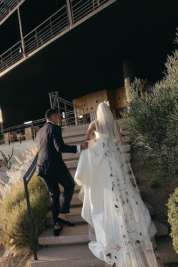 Wedding couple portrait of bride and groom walking up outdoor stairs, groom helping with dress train as long embroidered veil flows behind