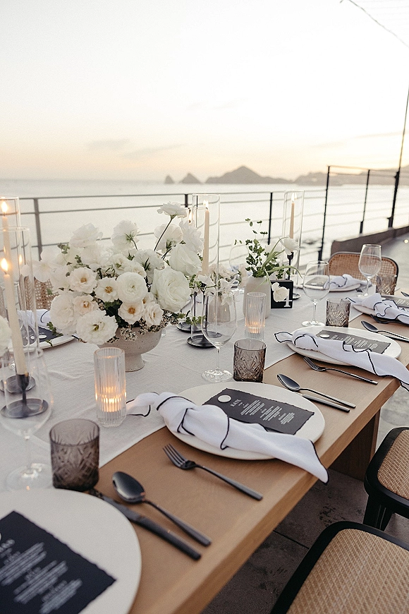 Reception tablescape with white floral centerpiece, taper and votive candles, black-and-white place settings on an oceanfront terrace at sunset