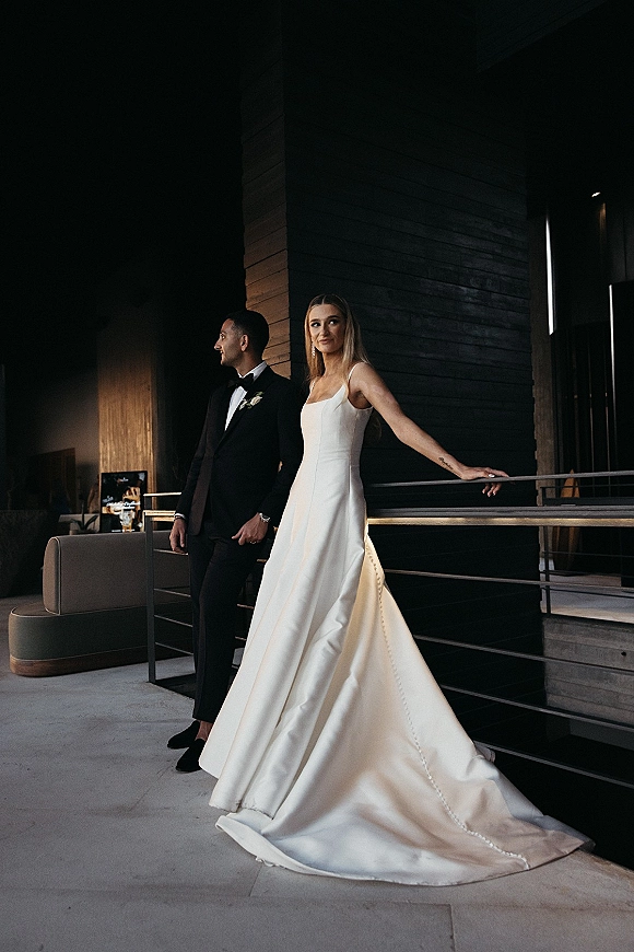 Couple portrait of bride and groom posing by a metal railing, her satin wedding dress and his black tuxedo lit by window light in a modern interior