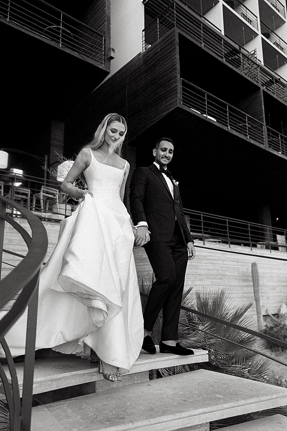 Wedding couple portrait in a black and white wedding photo, holding hands while walking down an outdoor staircase by a modern building with palm and cactus.