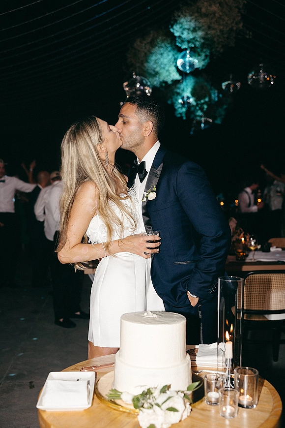 Wedding kiss portrait of newlyweds kiss at reception beside a simple white cake, bride in mini dress holding a cocktail glass under disco balls