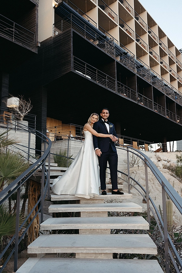 Couple portrait of bride in satin wedding dress hugging groom in black tuxedo on an outdoor staircase by a modern building and desert plants