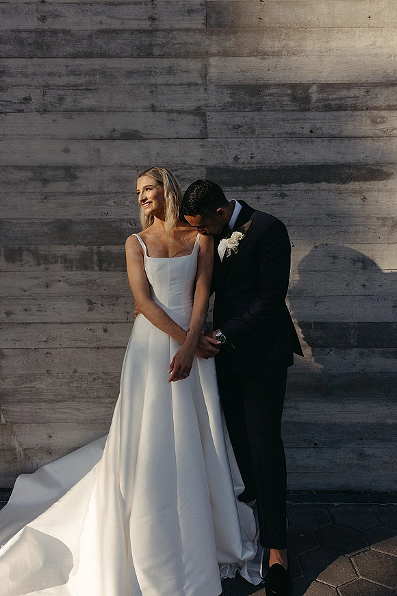 Couple portrait of bride and groom portrait, groom hugging bride as she looks away in a strapless gown by a wood plank wall backdrop