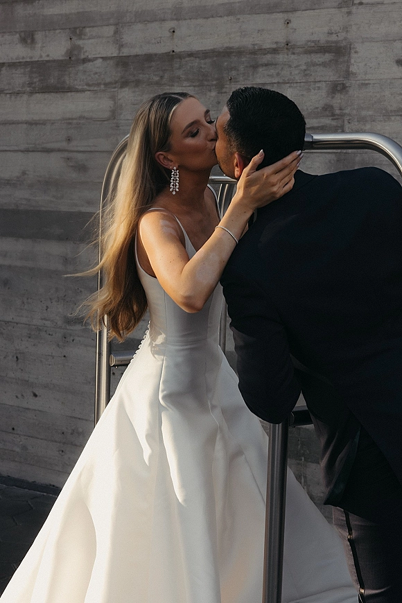 Wedding kiss portrait of bride and groom kissing, bride holding his face in strapless dress and drop earrings against a concrete wall
