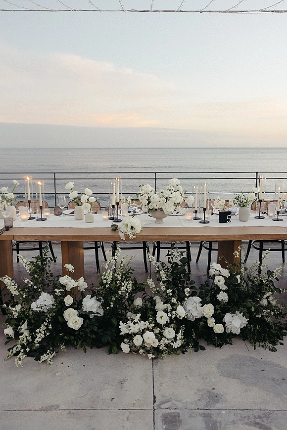 Reception tablescape with long head table decor featuring white and green florals, taper candles, and glassware on an oceanview terrace under string lights