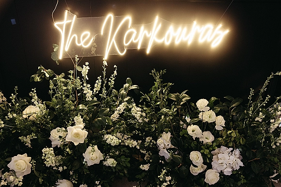 Wedding neon sign with custom last name neon glowing warm white on acrylic, framed by greenery and white roses against black drape backdrop