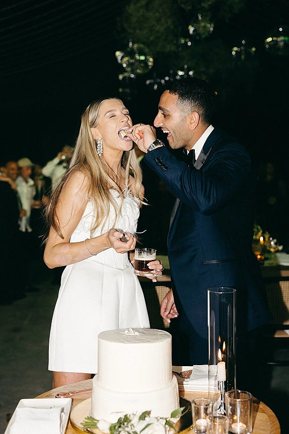 Cake cutting moment as bride in a short wedding dress and groom in a tux slice a two-tier white cake under disco balls at night reception