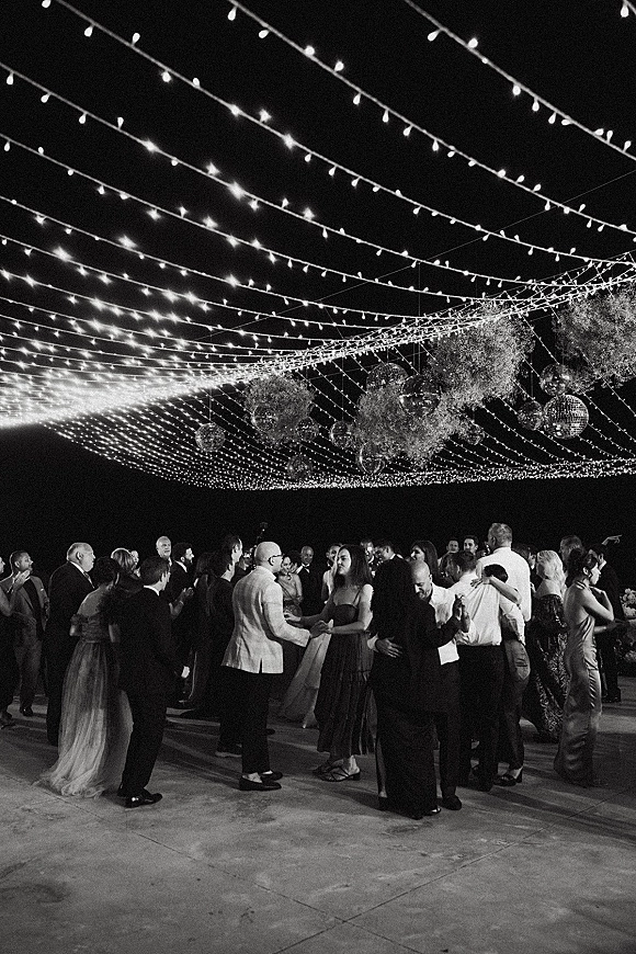 Wedding dance floor packed with guests dancing under string lights, with disco balls and hanging florals on an outdoor patio at night