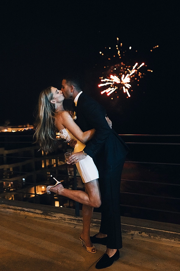 Wedding kiss portrait of newlyweds embracing on a railing, bride in a white mini dress holding a drink as fireworks burst over city lights waterfront at night