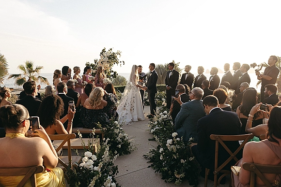 Wedding ceremony with bride and groom under a white floral arch, oceanfront terrace at sunset with palm trees and guests seated