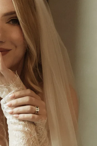 Bridal portrait of a bride with veil covering her face, showing lace sleeve cuff, nude manicure, and engagement ring on a neutral wall