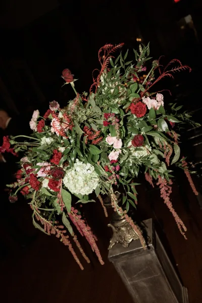 Wedding floral arrangement in an ornate pedestal vase with red and blush roses, hydrangea, and greenery against dark candlelit bokeh background