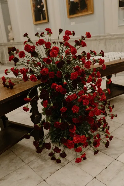 Ceremony aisle flowers in a wedding aisle floral arrangement of red carnations beside wooden pews in an indoor hall with a stone wall