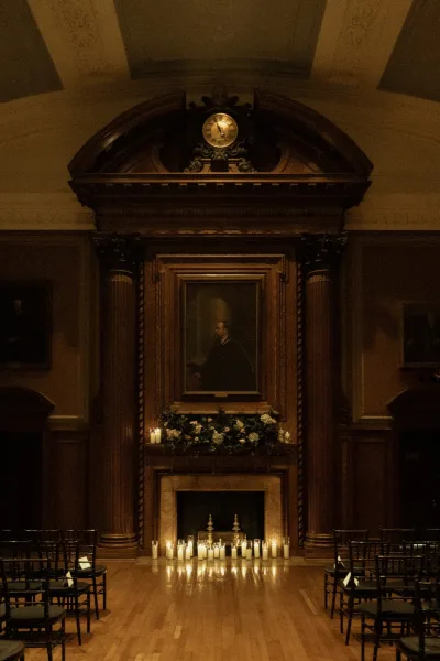 Indoor ceremony setup with a candlelit wedding ceremony by a grand fireplace altar, black chairs and pillar candles in a wood-paneled room